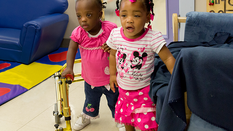 Two toddlers are standing near couches in a childcare setting. One is leaning on a walker.