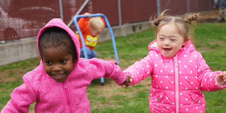 Preschoolers wearing hoodies and jackets, play outside. Two girls run holding hands in the foreground while a boy plays on an individual trampoline in the background. 